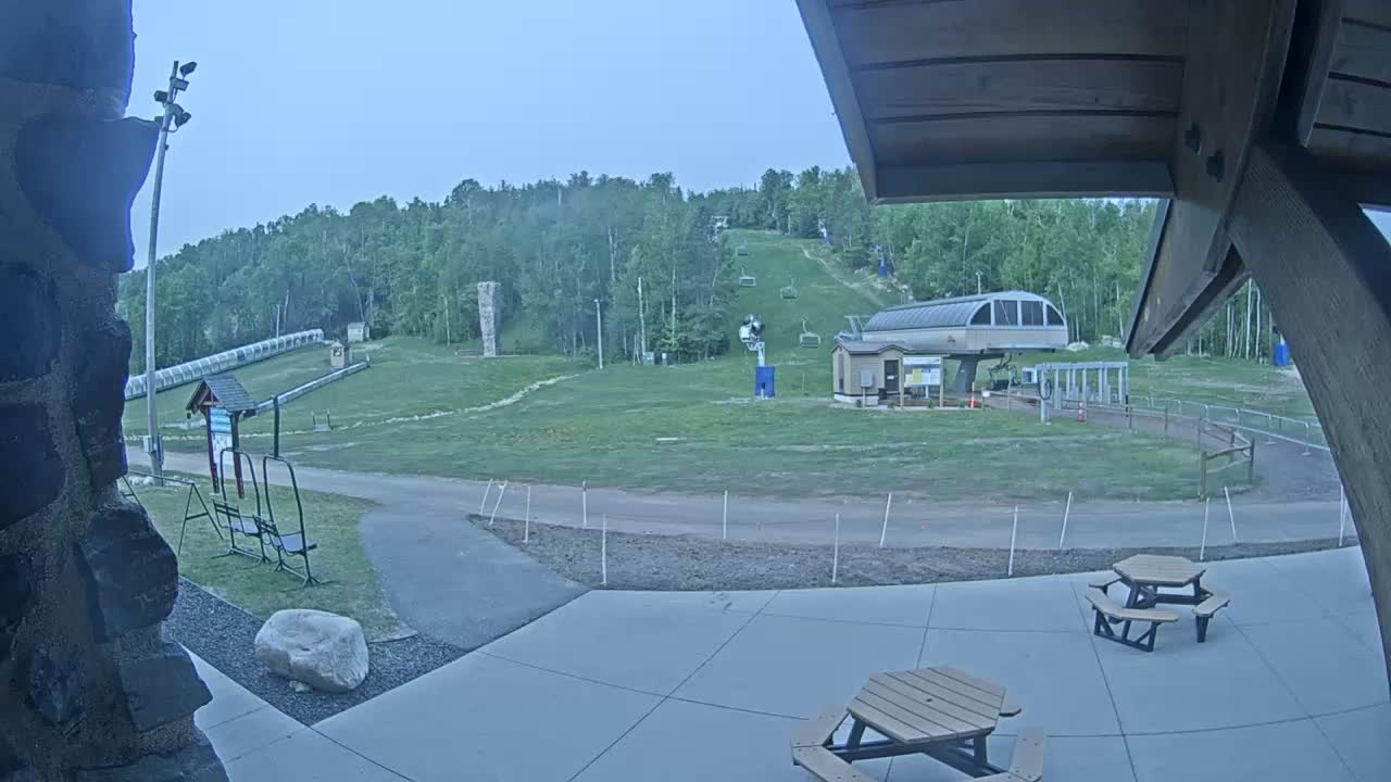 A ski resort's base area, under an overcast sky, shows a chairlift, a grassy hill with ski runs, picnic tables, and a paved patio.