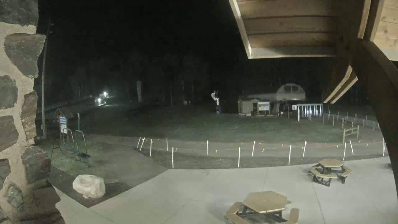 A nighttime view from under a wooden structure shows a paved area with picnic tables, a path bordered by small markers, and a distant building under dark, nighttime conditions.