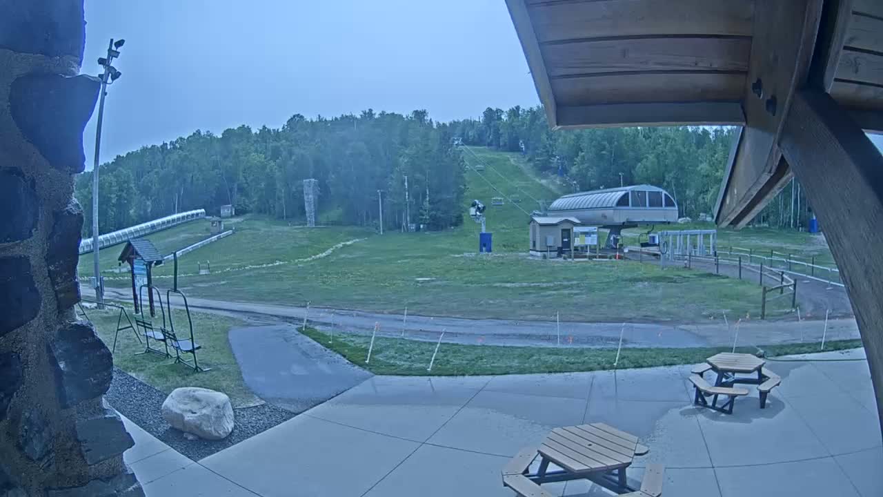 A ski lift and grassy hill are visible under a mostly cloudy sky, with picnic tables and a paved area in the foreground.