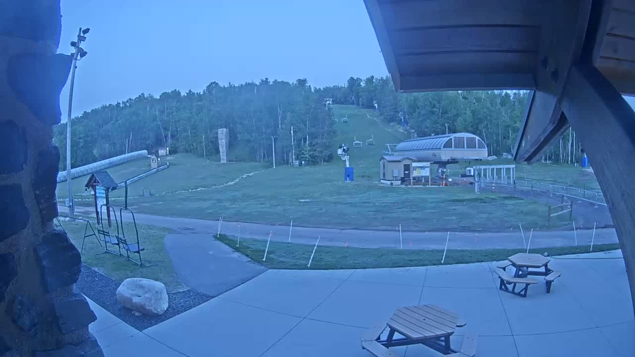 An outdoor scene at dusk shows a ski lift and hill, along with picnic tables and other outdoor furniture in a grassy area.