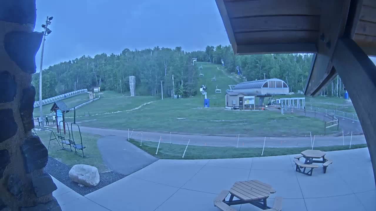 A ski lift and slope are visible in the distance under a twilight sky, with picnic tables and a paved area in the foreground.