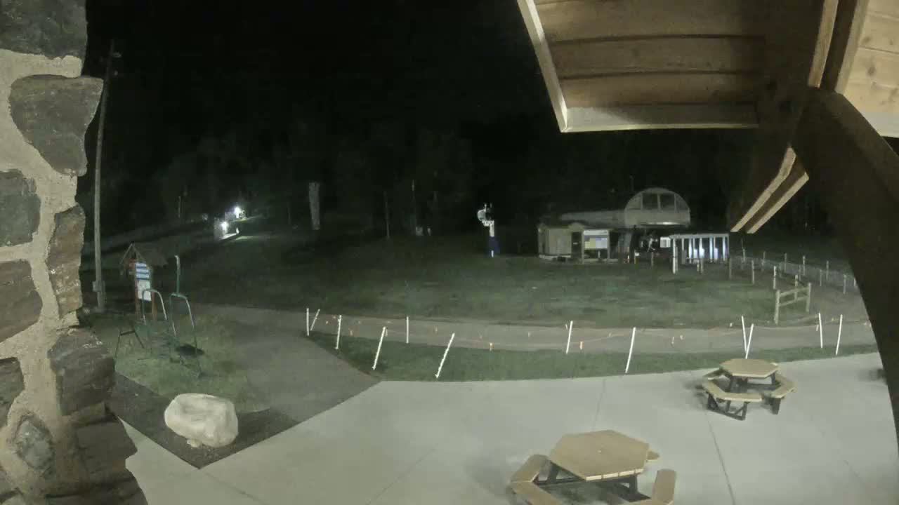 A nighttime view of a paved area with picnic tables, a grassy field with some structures and a path, and a stone wall.