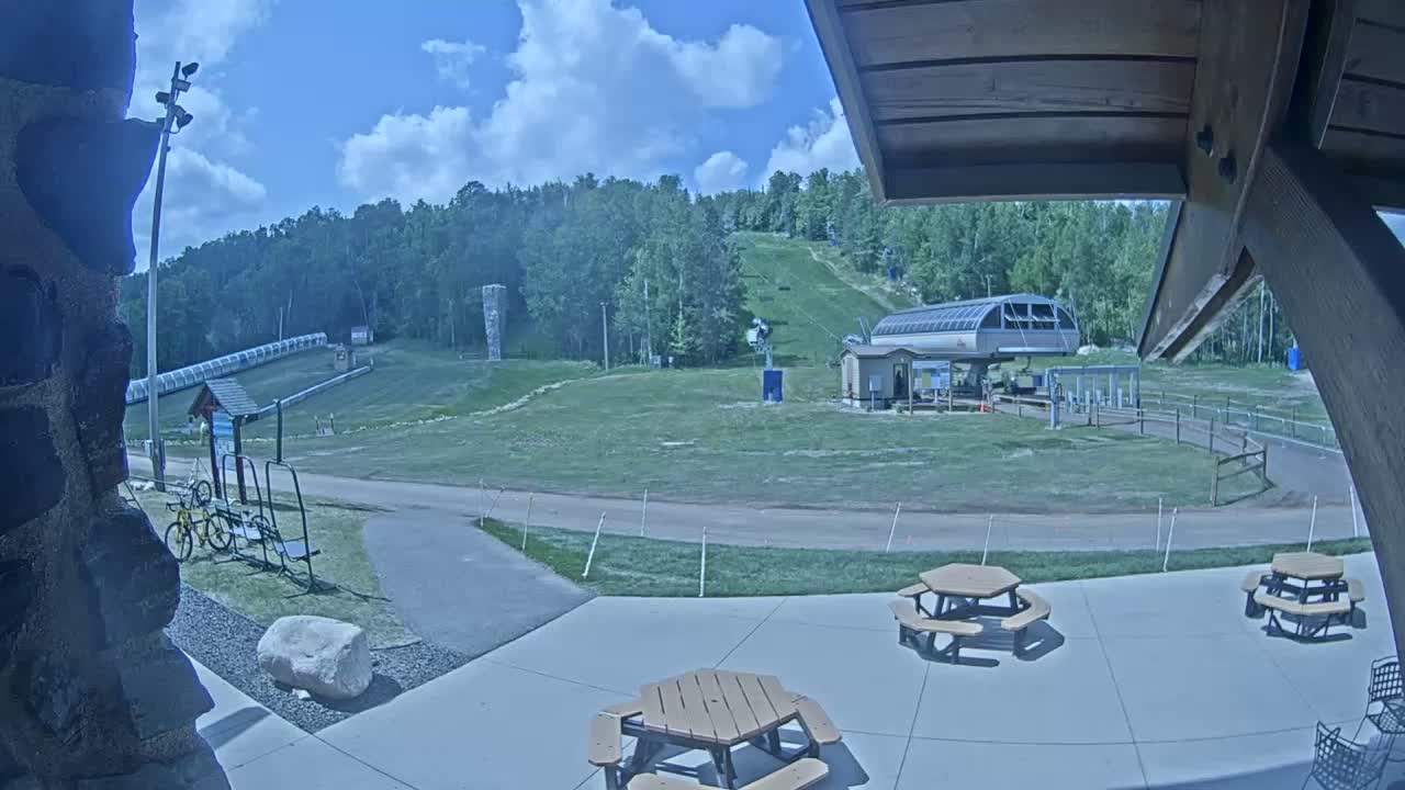 A sunny outdoor scene shows a ski lift and grassy hill, with picnic tables on a patio in the foreground and a bicycle leaning against a rack.