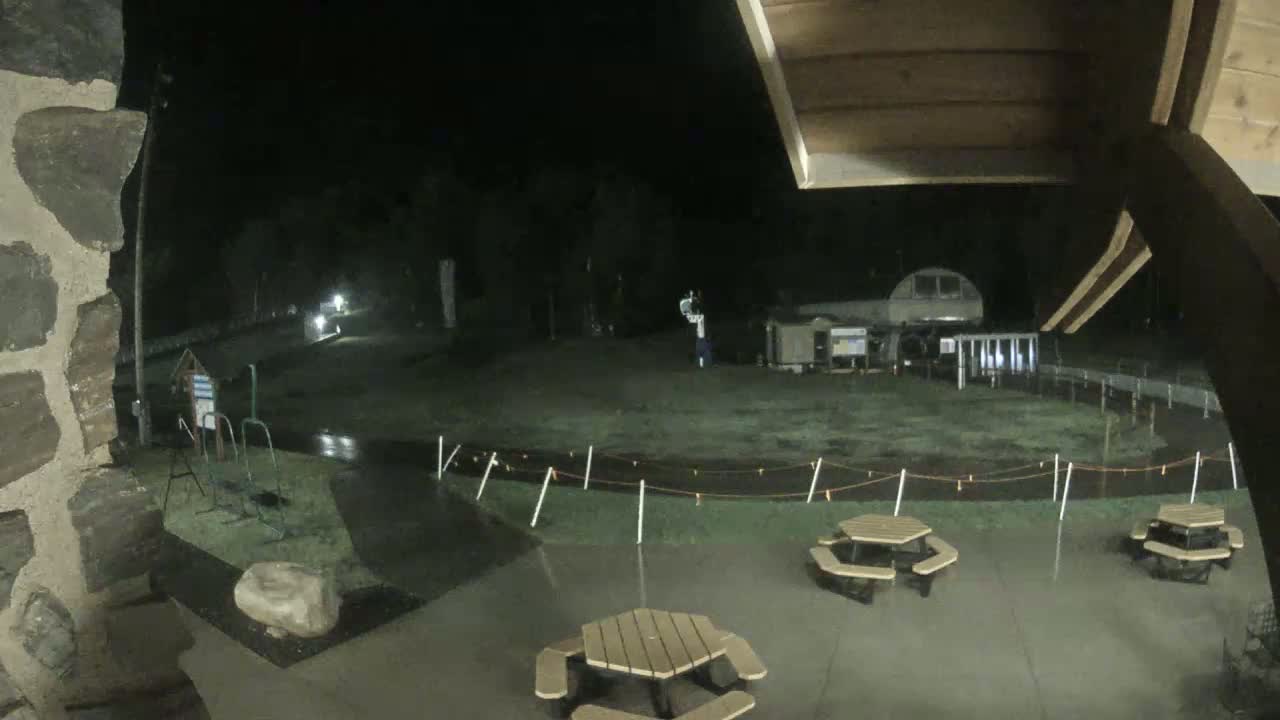 At night, under rainy conditions, a wet outdoor area with picnic tables is seen from under a covered structure, with a building and some equipment visible in the background.