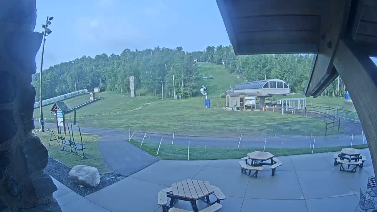 A view from a patio shows a grassy area with a ski lift and a forested hillside in the background on a mostly clear day.