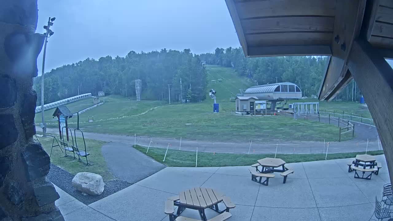 An overcast outdoor scene shows a grassy ski slope with a chairlift and picnic tables on a patio in the foreground.