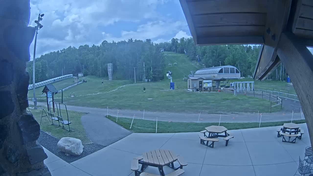 An outdoor patio area with picnic tables overlooks a grassy ski slope with a chairlift and a covered conveyor lift under a partly cloudy sky.