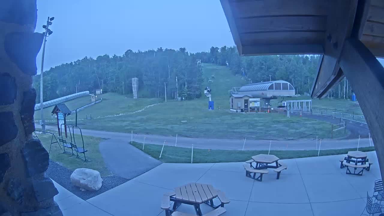 An outdoor scene at dusk shows a grassy ski slope with a chairlift and a small building, picnic tables on a patio in the foreground.