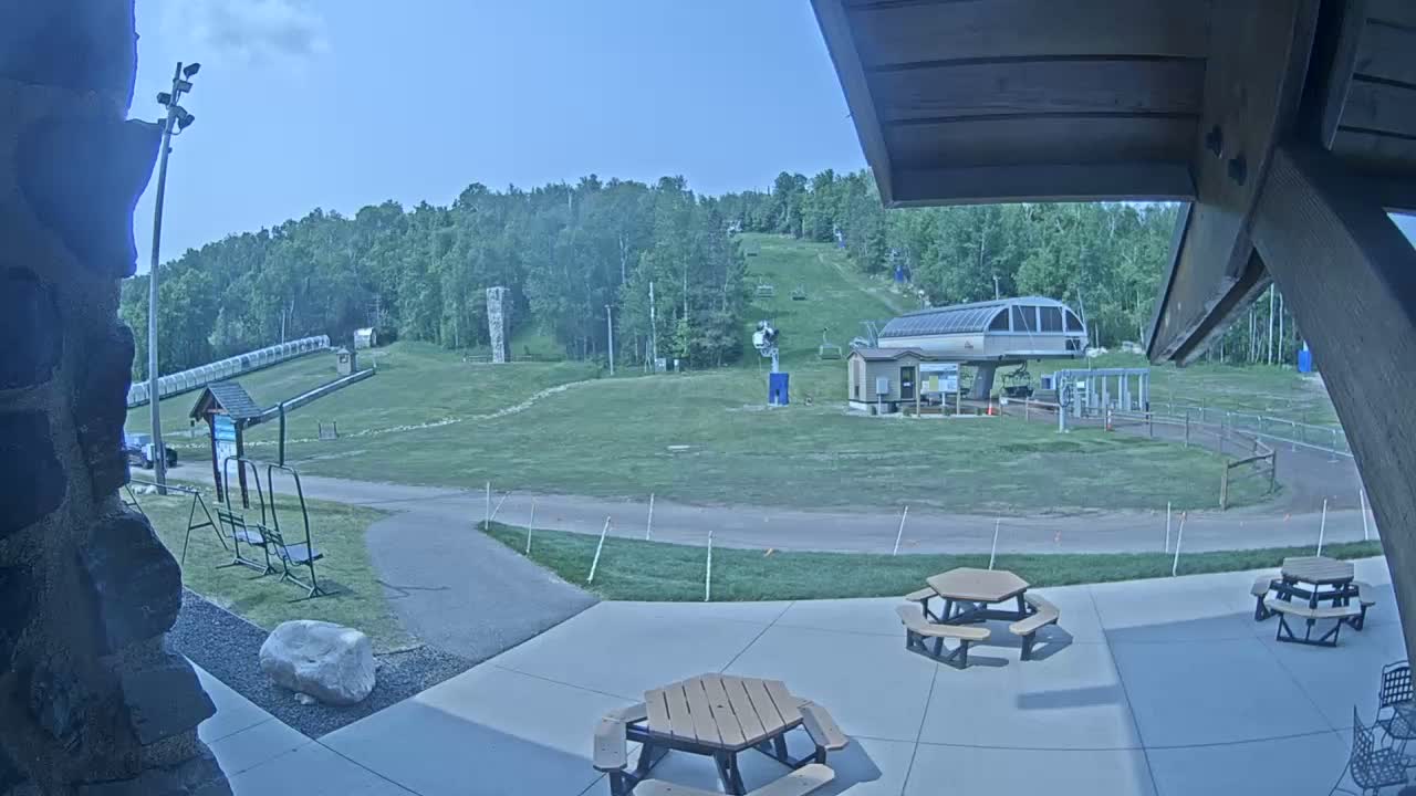 A grassy ski slope with a chairlift and several picnic tables on a patio in sunny weather.