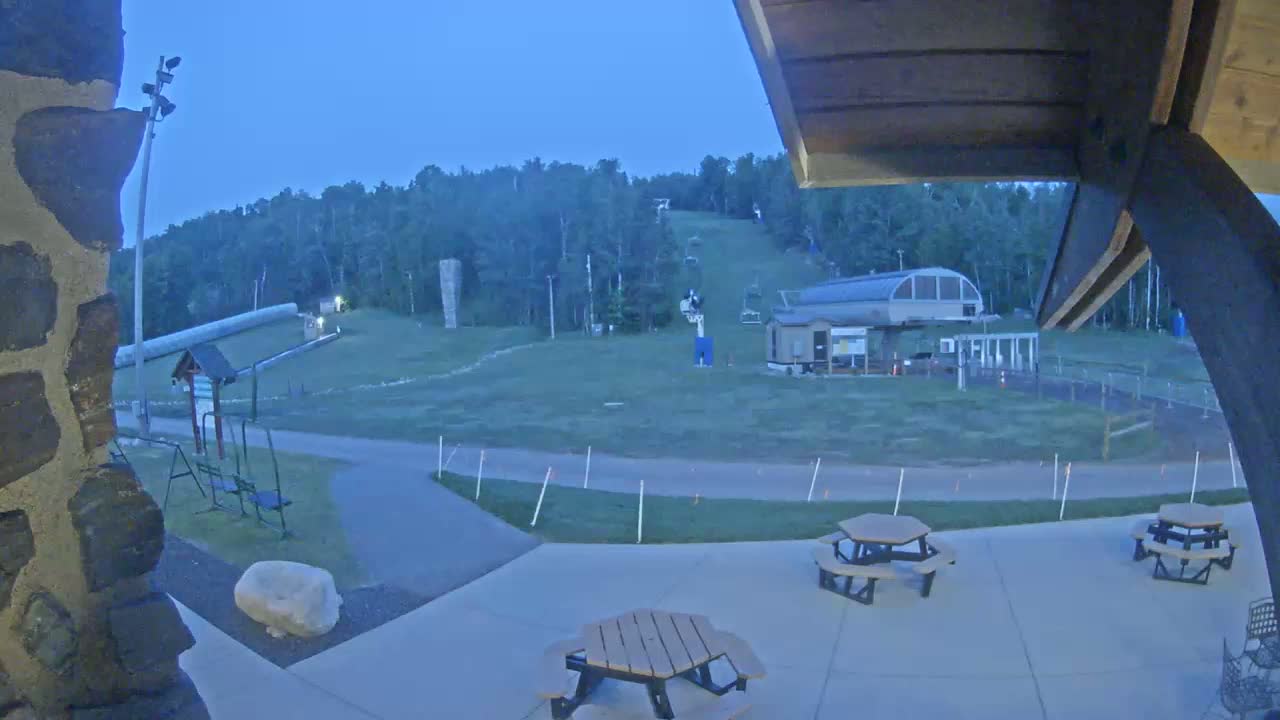 A nighttime view of a grassy ski slope with a chairlift, a small building, picnic tables, and a stone wall under a twilight sky.