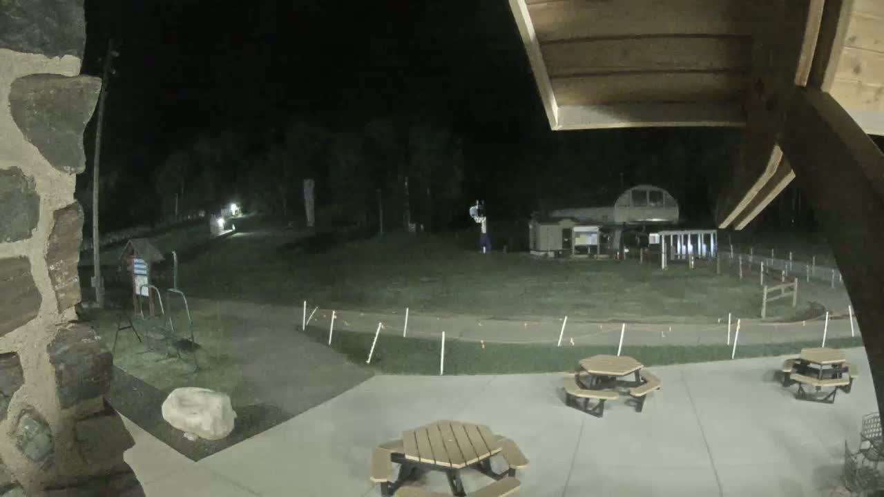 At night, a paved area with hexagonal picnic tables is visible, overlooking a grassy field with a small building and playground equipment under dim lighting.