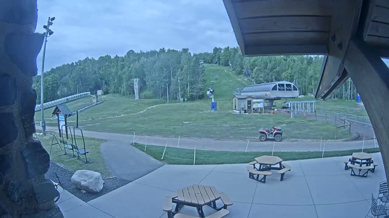 An outdoor patio with picnic tables overlooks a grassy ski slope with a chairlift and an ATV, under an overcast sky.