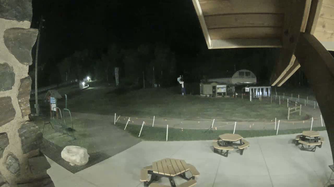 At night, a paved area with picnic tables is seen from under a wooden overhang, overlooking a grassy field with a distant structure and some small lights.