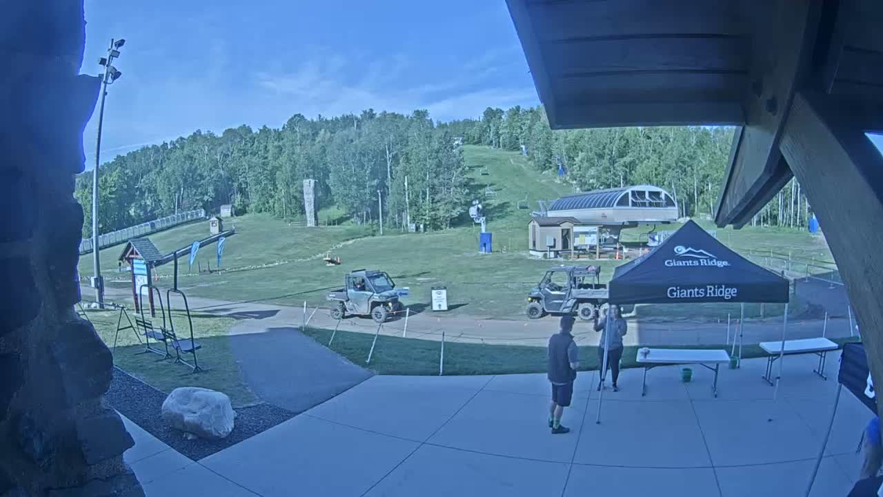 A sunny outdoor scene shows a grassy ski hill with two utility vehicles parked near a small building, a canopy tent, and two people standing on a paved area.