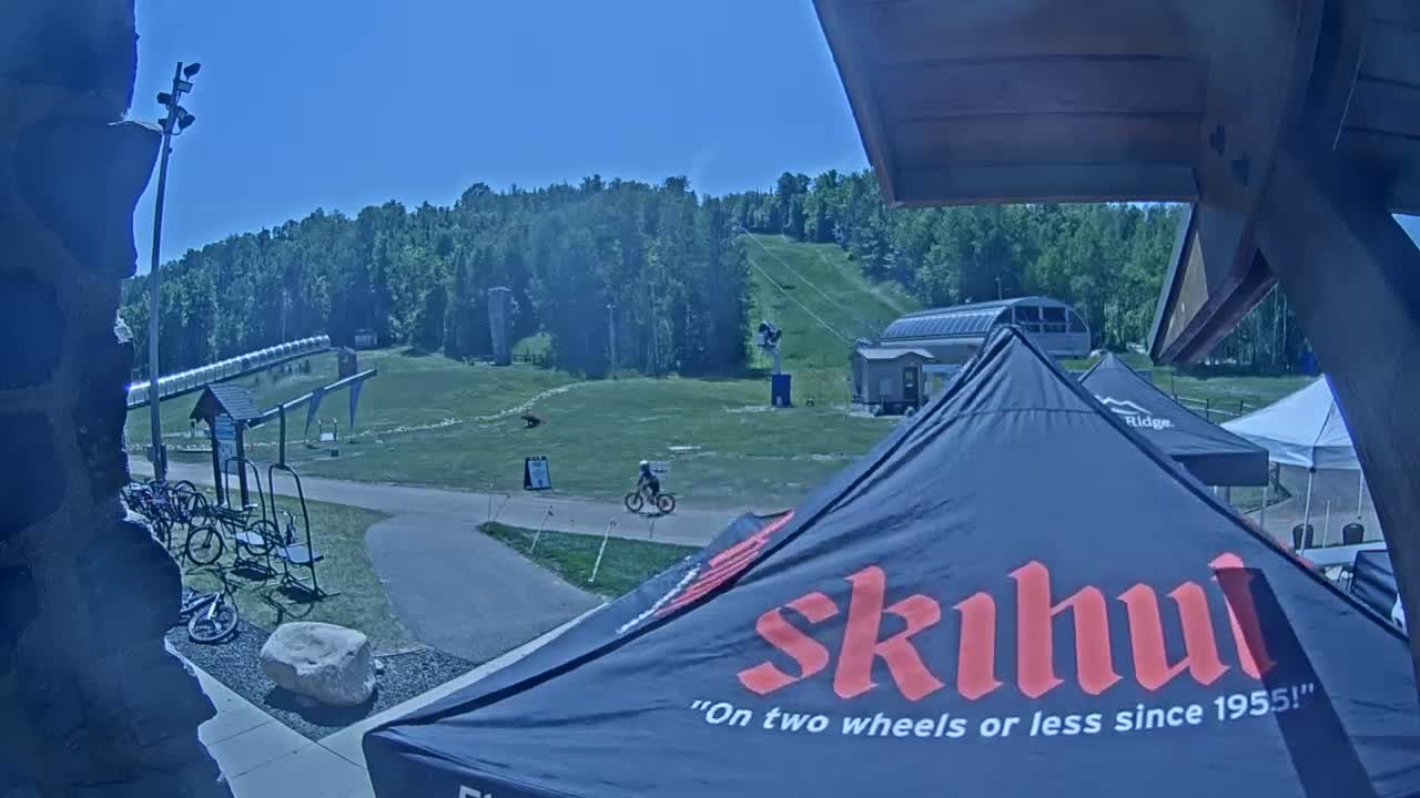 A sunny outdoor scene shows a grassy ski hill with two utility vehicles parked near a small building, a canopy tent, and two people standing on a paved area.