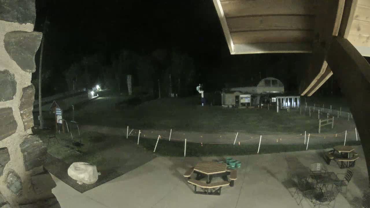 A nighttime outdoor scene shows a paved area with picnic tables, a stone wall, and a grassy field beyond with some buildings and fencing in the distance under a dark sky.