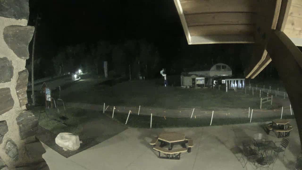 A nighttime view of an outdoor area with picnic tables, a fenced-off grassy field, and a building in the background.