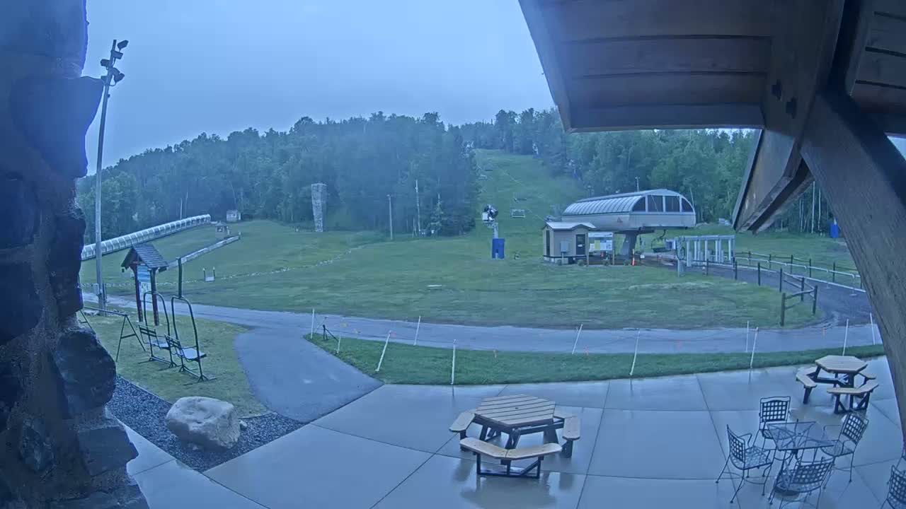 A wet patio with picnic tables and chairs overlooks a grassy ski slope with a chairlift and a long, low structure under an overcast sky.