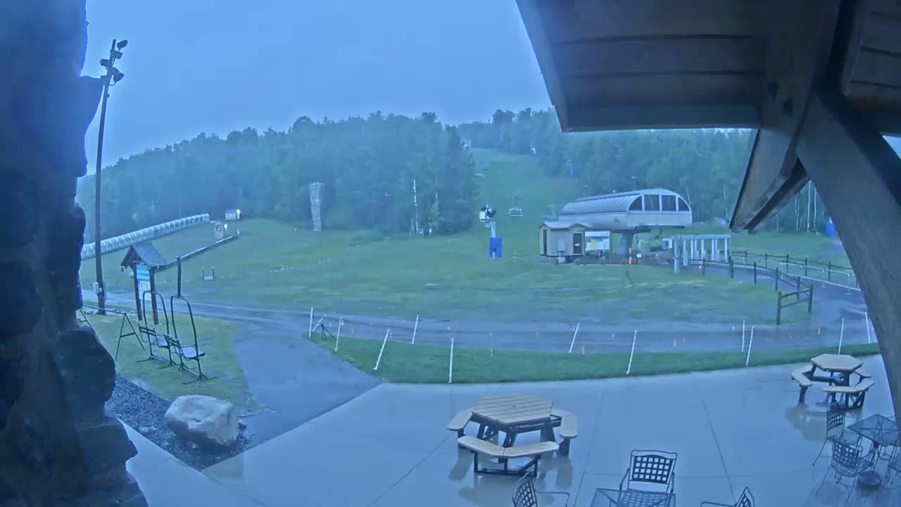 A rainy view of a ski resort's base area with a chairlift, picnic tables, and a grassy slope in the background.