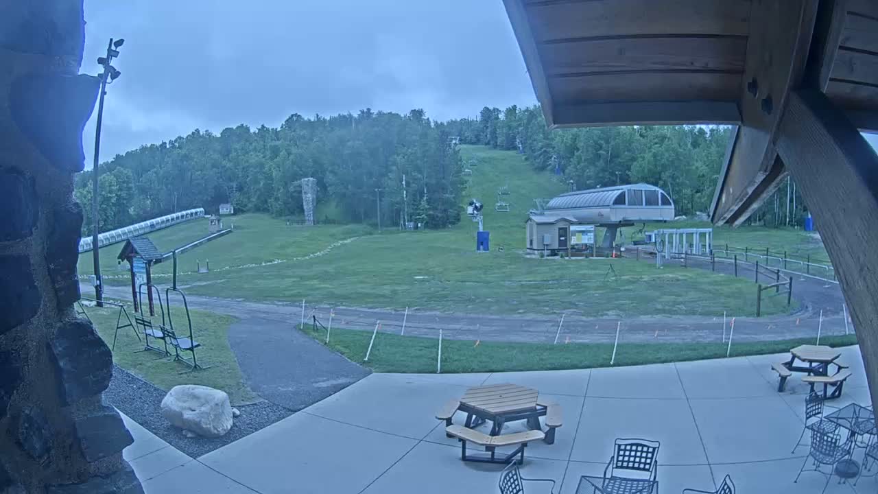 A grassy ski slope with a chairlift and other ski infrastructure is visible from a patio with picnic tables under an overcast sky.
