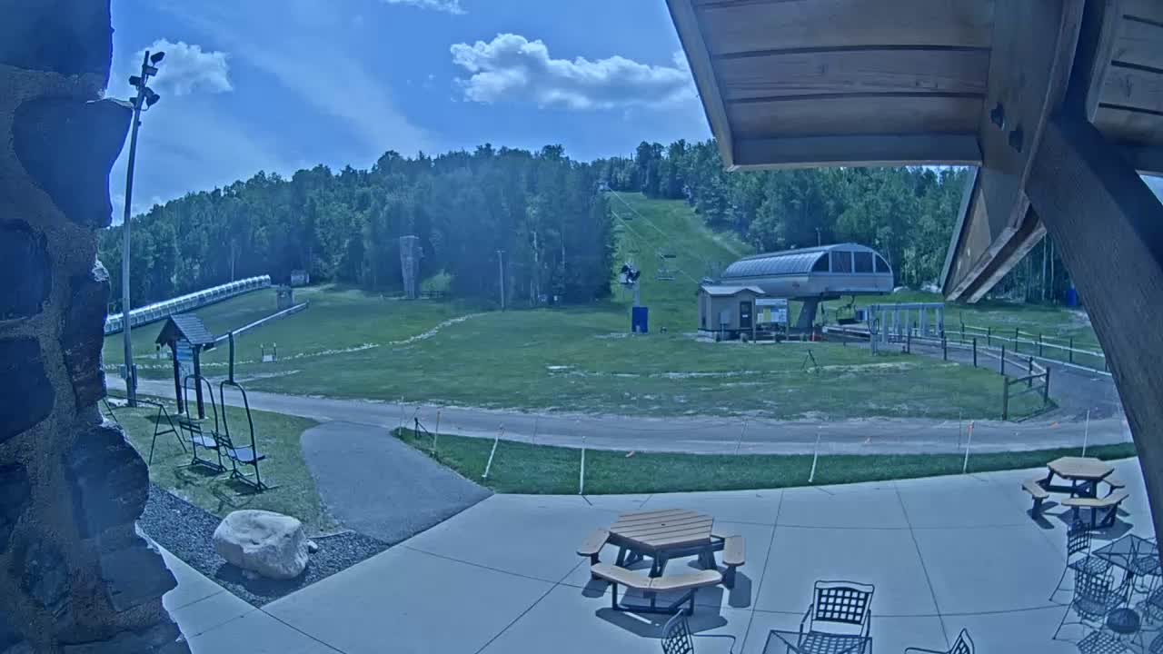 A mostly sunny outdoor scene shows a ski lift and grassy slopes, with picnic tables and chairs on a patio in the foreground.