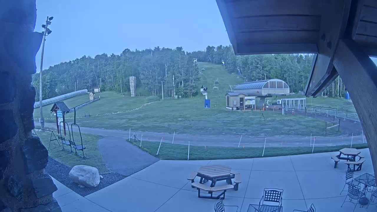 A grassy ski slope with a chairlift and small building is visible from a patio with picnic tables under a twilight sky.
