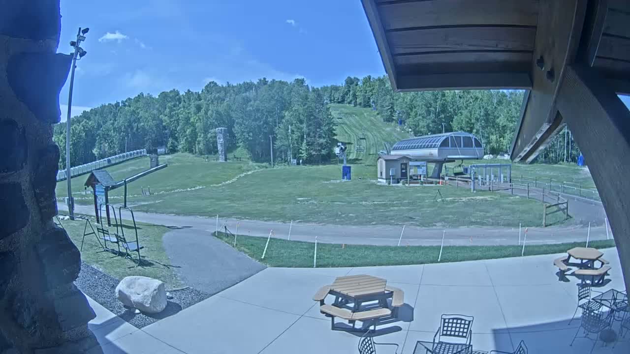 A mostly sunny outdoor patio area with picnic tables and chairs overlooks a grassy ski slope with a chairlift in the background.