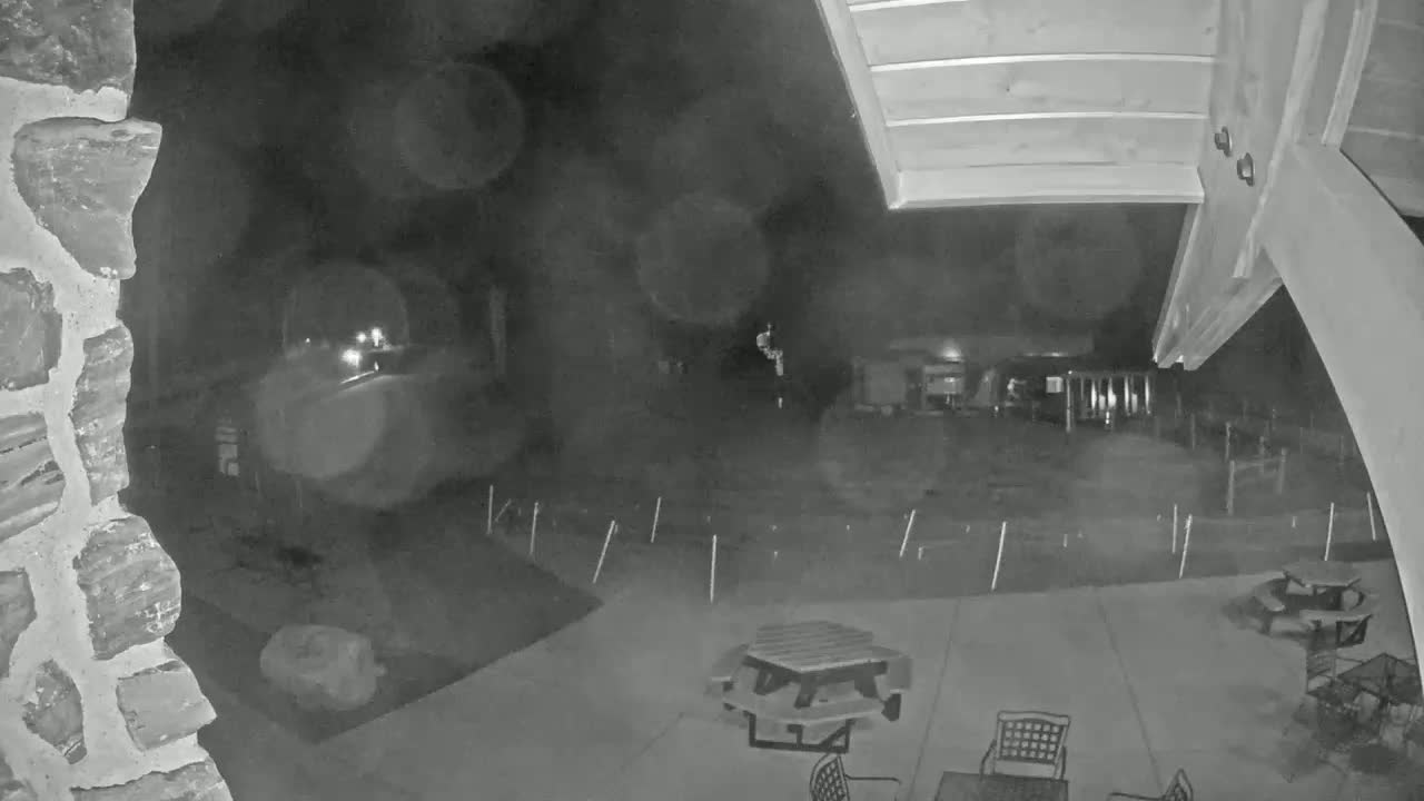 A nighttime, blurry, low-light security camera view shows a patio area with picnic tables and chairs, a stone wall to the left, and a dimly lit yard extending to the background.