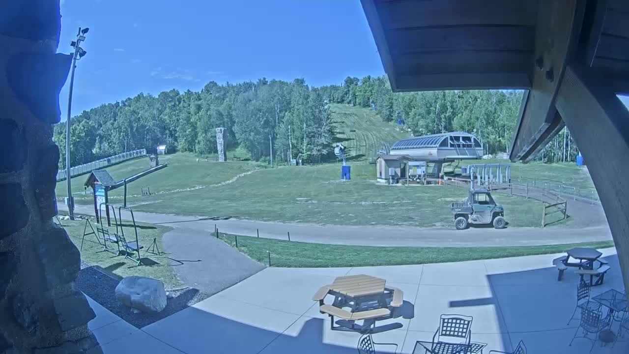 A sunny outdoor patio area with picnic tables and chairs overlooks a grassy ski slope with a chairlift and utility vehicle in the distance, surrounded by a forest.