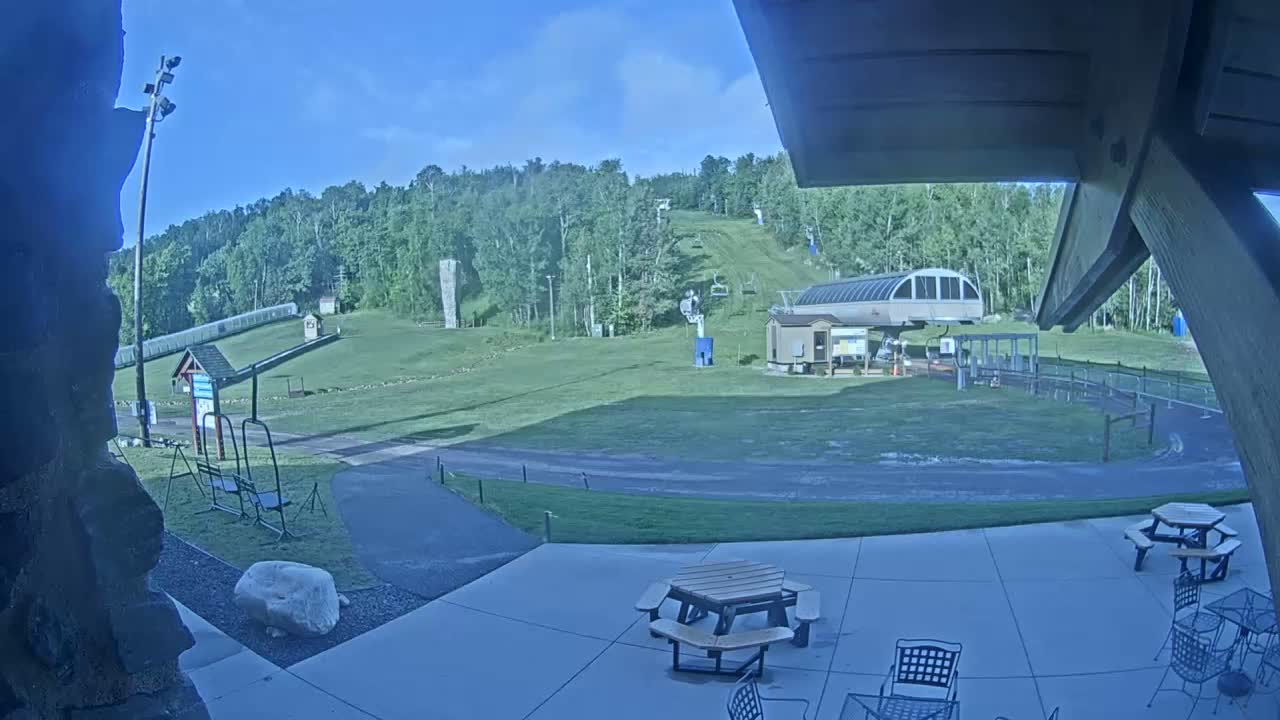 A patio with picnic tables overlooks a grassy ski slope with a chairlift and surrounding trees under a clear blue sky.