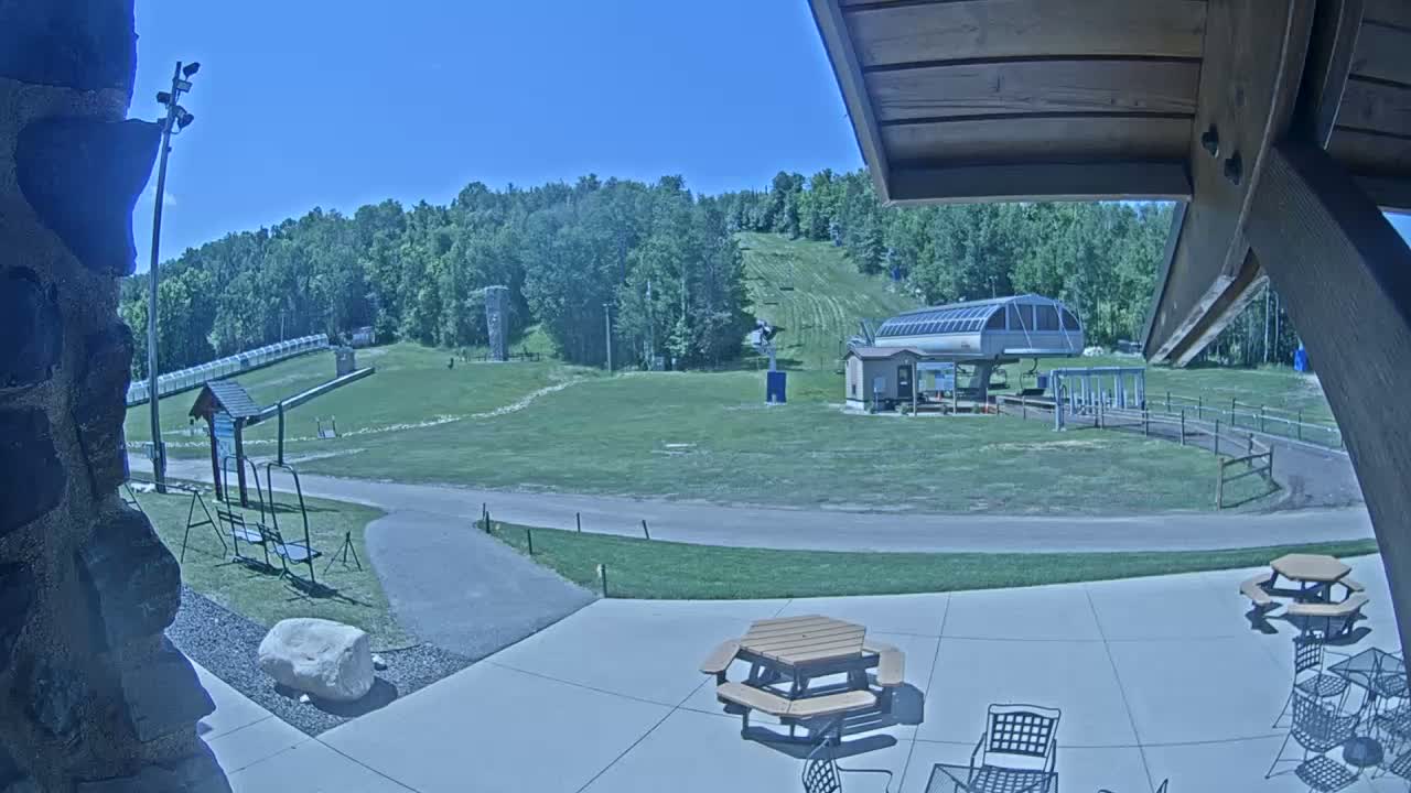 A sunny outdoor scene shows a ski lift and grassy hills in the background, with picnic tables and chairs on a patio in the foreground.