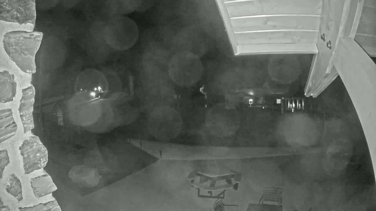 A nighttime, blurry outdoor security camera view shows a stone wall, a partially visible picnic table, and a dark yard with indistinct features beyond.