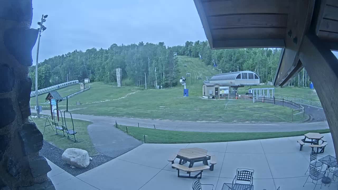 A grassy ski slope with a chairlift and a covered patio area with picnic tables, viewed on an overcast day.