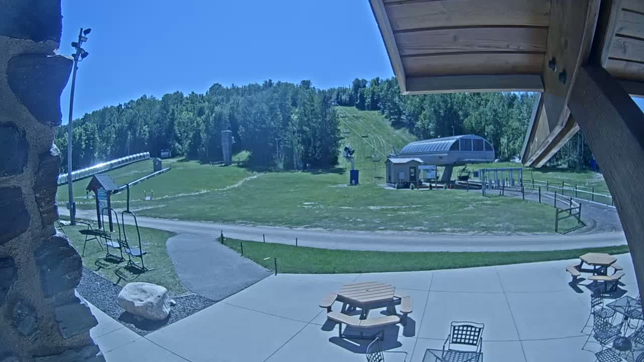 A sunny outdoor scene shows a ski lift and grassy hill with picnic tables and chairs on a patio in the foreground.