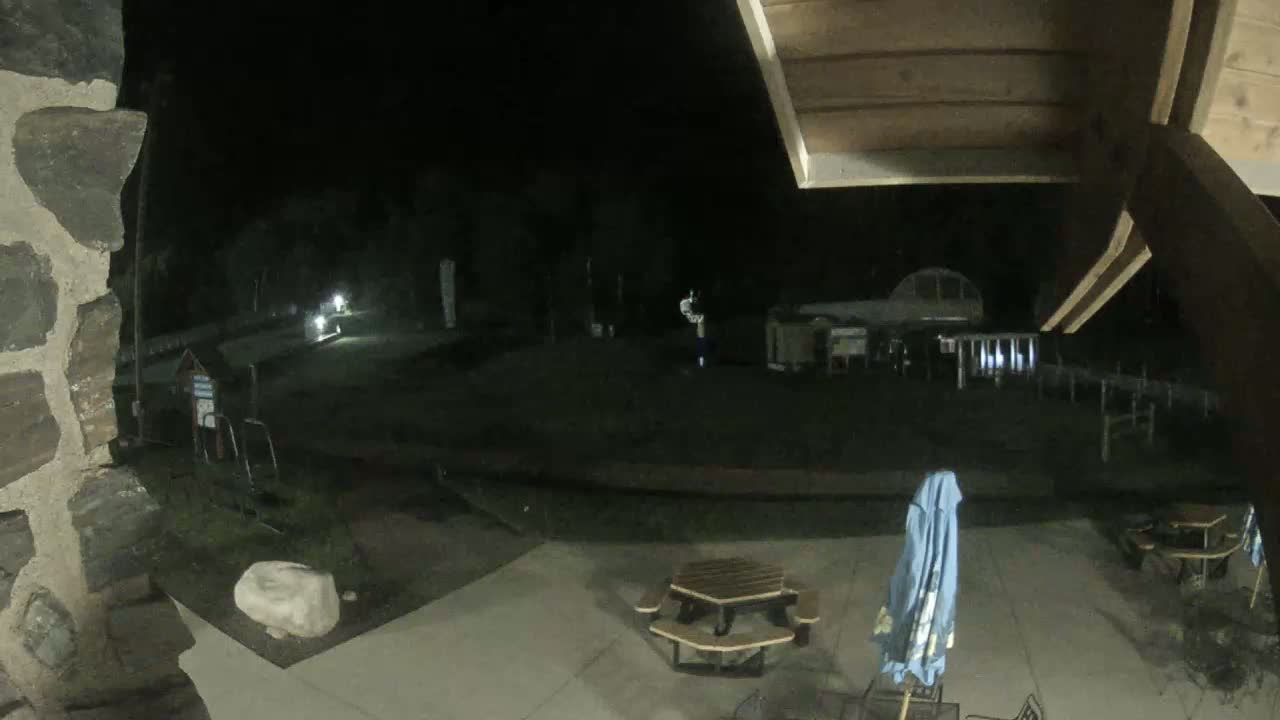 A nighttime view from under a wooden structure shows a paved area with picnic tables, a folded beach umbrella, and a dimly lit outdoor space beyond.