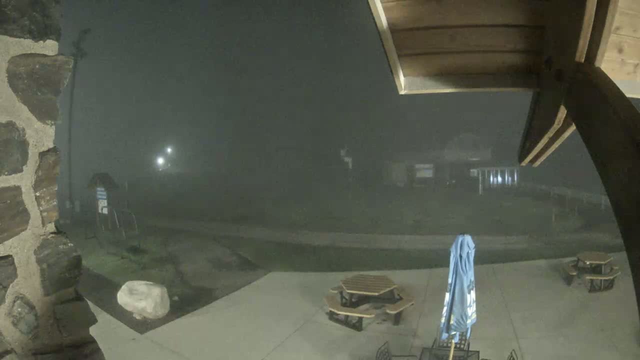 A nighttime, foggy view shows a stone wall, patio furniture, and a closed umbrella on a concrete patio, with a building faintly visible in the background.