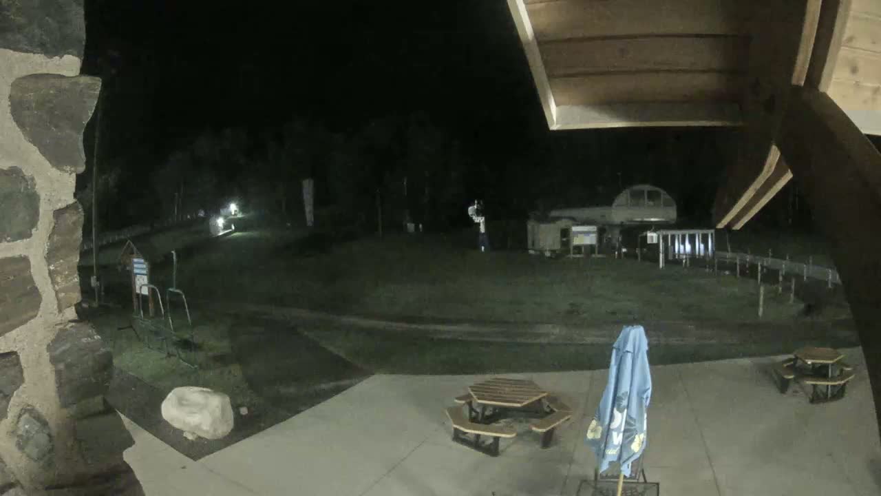 A nighttime view of a paved area with picnic tables and a closed umbrella, overlooking a grassy field with a distant building and some playground equipment under a dark sky.