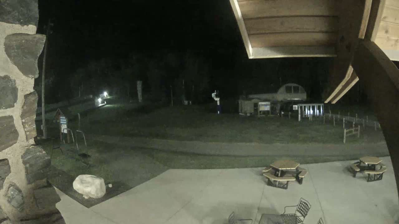 A nighttime outdoor scene shows a partially visible stone structure, a grassy area with a building in the background, and a closed umbrella on a patio.