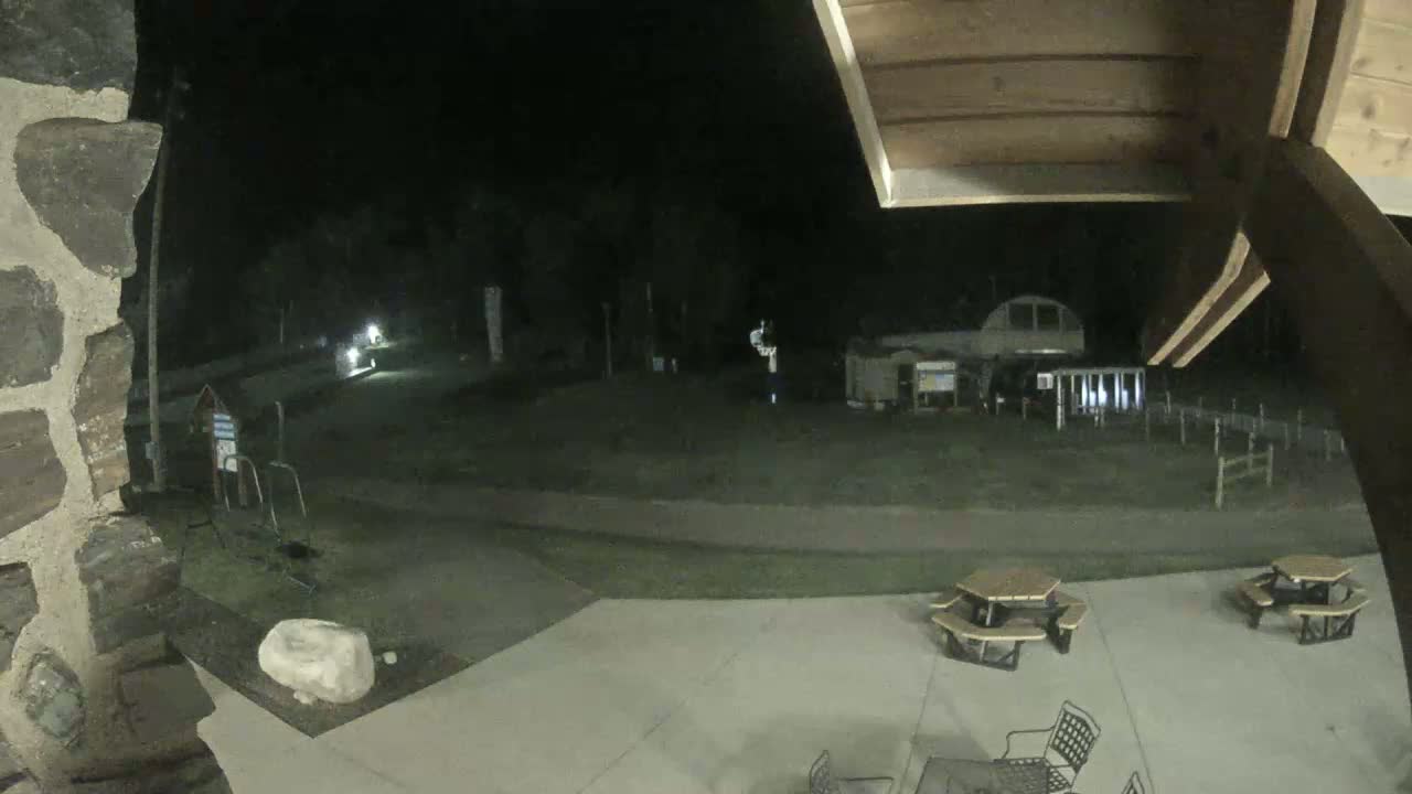 A nighttime outdoor scene shows a partially visible stone structure, a grassy area with a building in the background, and a closed umbrella on a patio.
