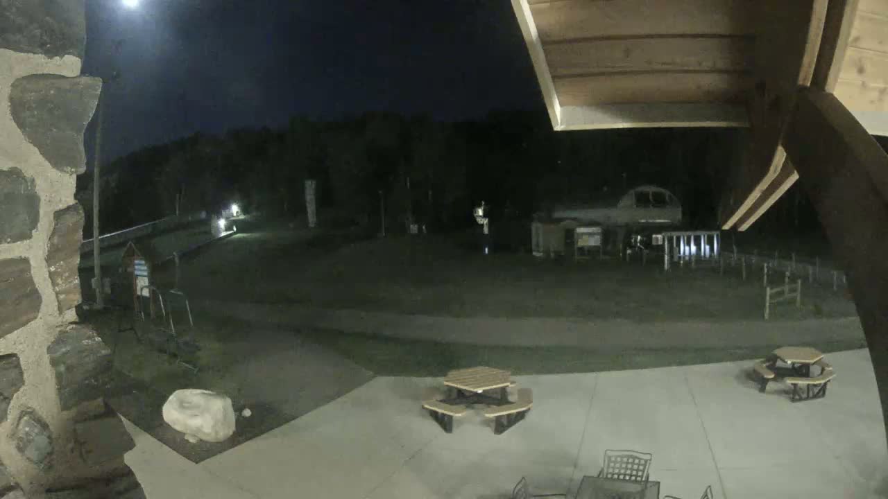 A nighttime view shows a paved area with picnic tables, a stone wall, and a vehicle parked in the background under a dark sky.
