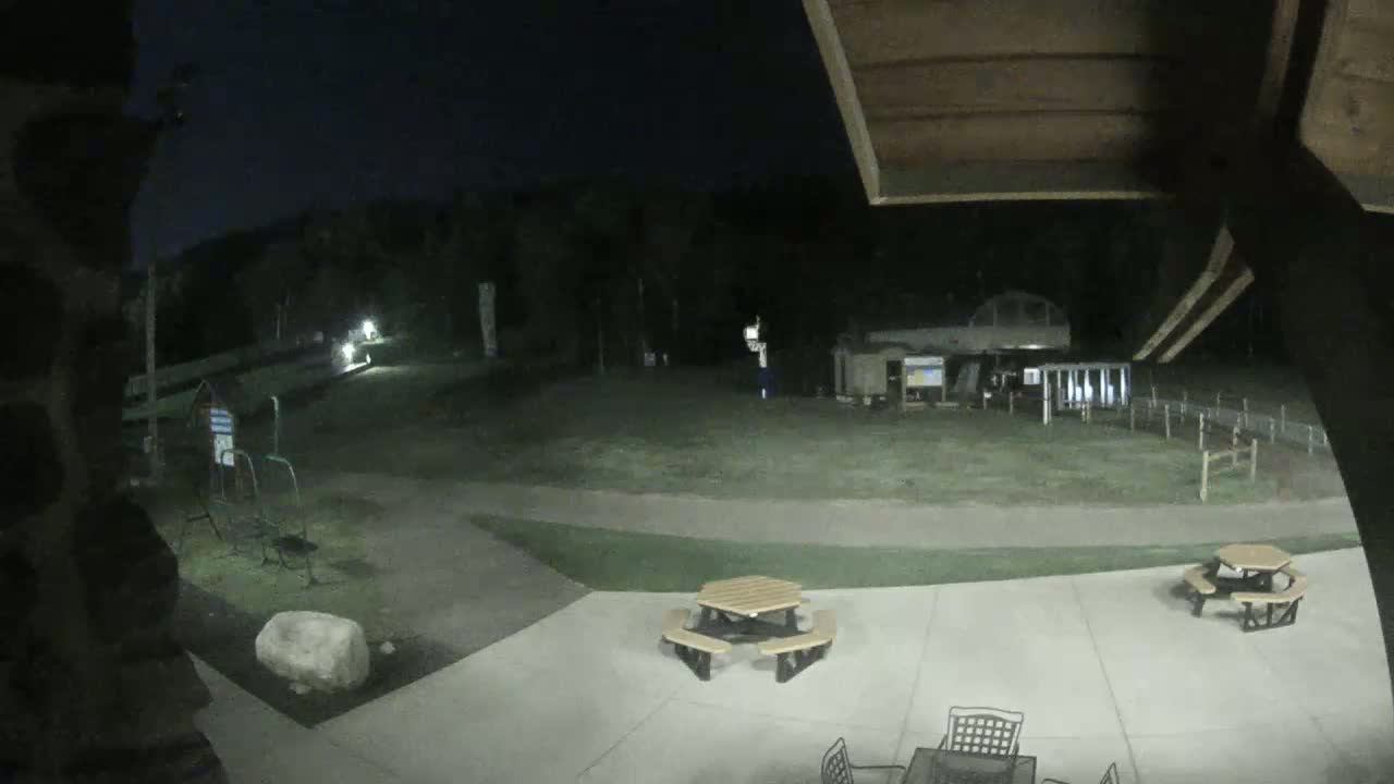 A nighttime view of a paved patio area with picnic tables, a grassy area beyond, and some structures in the background under a dark sky.