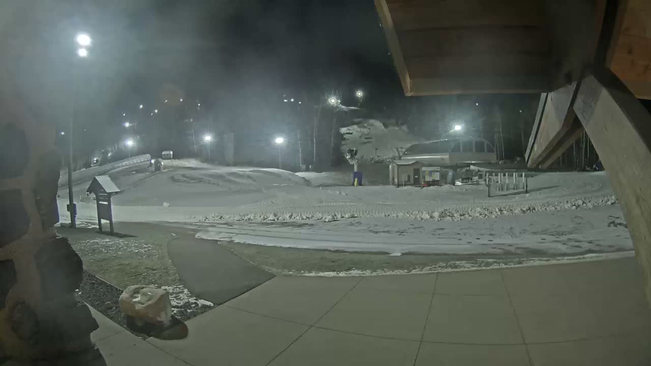 A nighttime view of a paved patio area with picnic tables, a grassy area beyond, and some structures in the background under a dark sky.