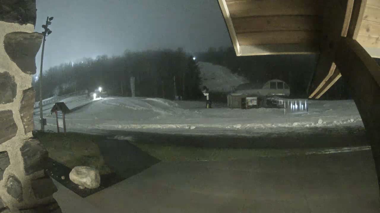 A misty or lightly snowing night at a ski resort reveals illuminated snow-covered slopes, a chairlift, and various buildings, framed by a stone wall on the left and a wooden roof structure on the right.