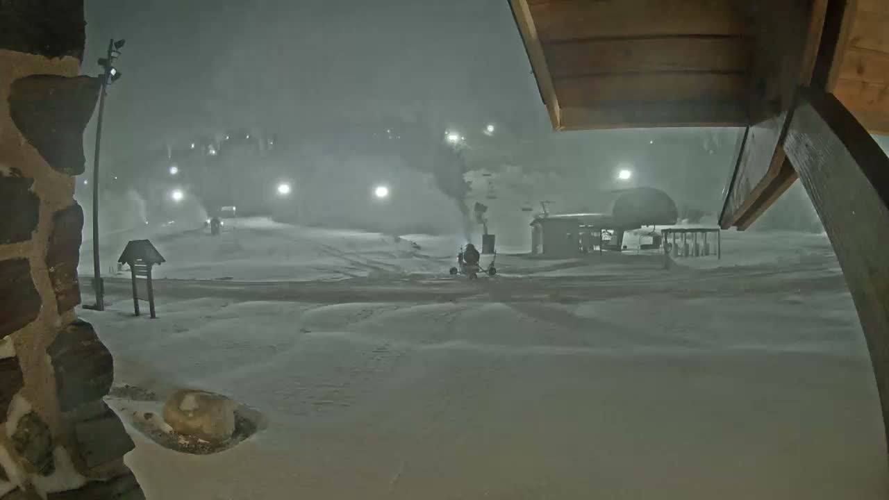 A snow-covered ski resort at night is illuminated by numerous diffused lights, with an active snowmaking machine spraying snow near a ski lift, suggesting cold and hazy conditions.