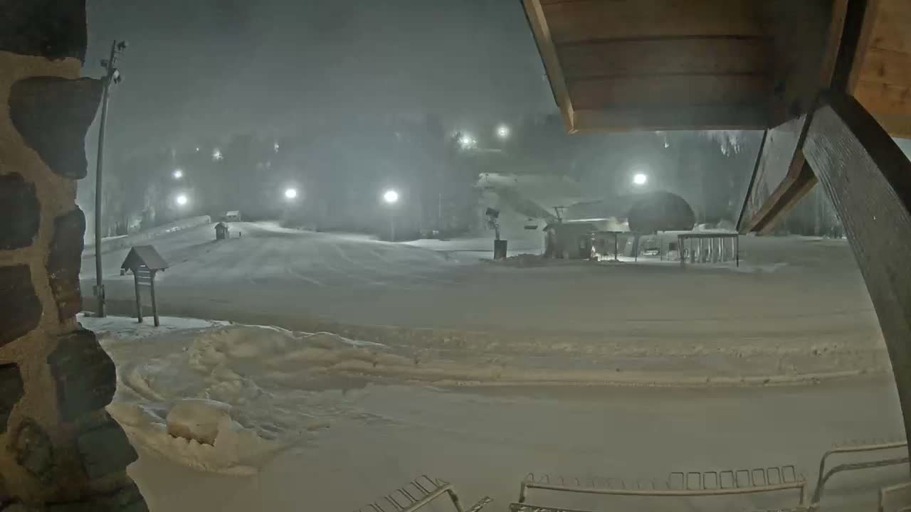 A snow-covered ski resort is brightly illuminated at night by numerous scattered lights, creating a hazy and foggy atmosphere over the slopes and facilities.
