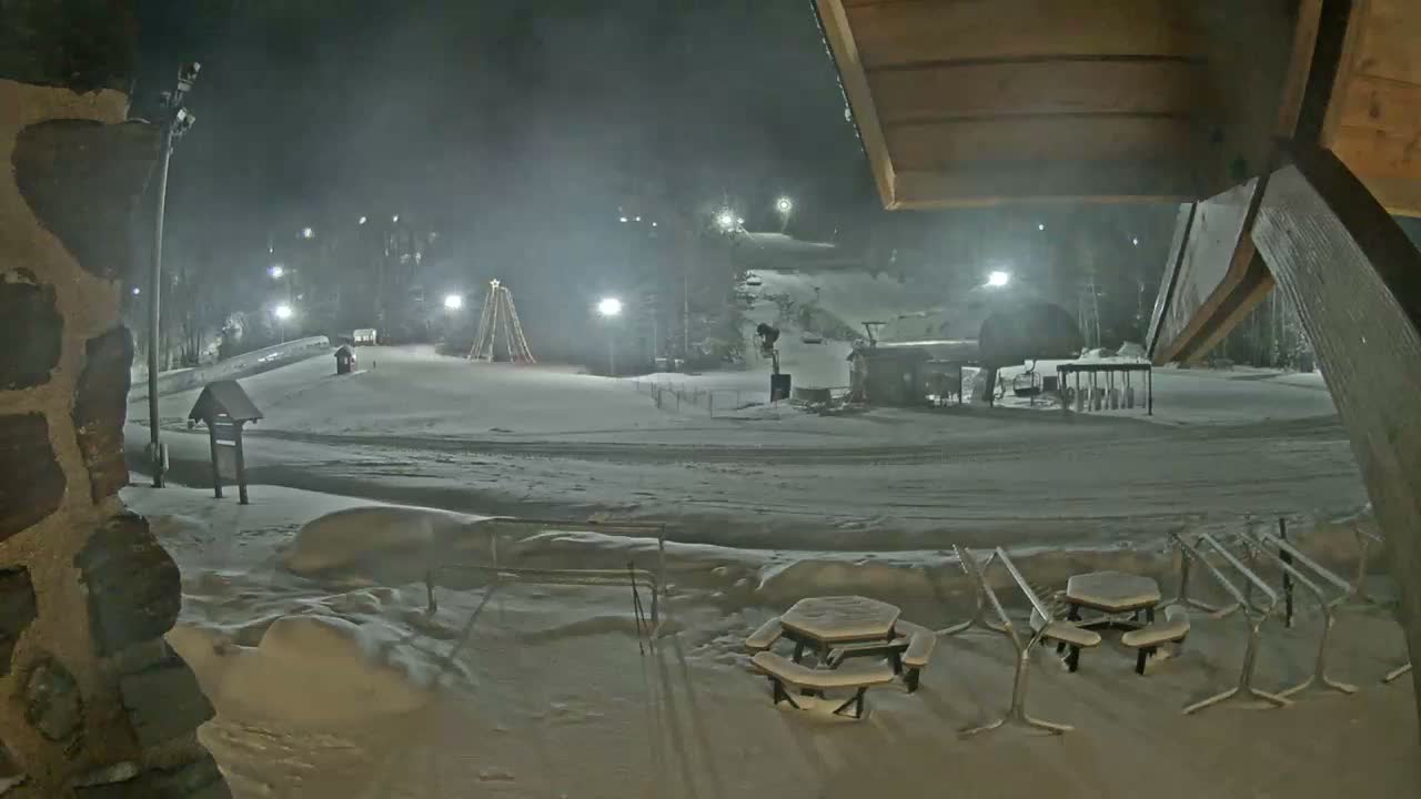 A snowy ski resort is illuminated by floodlights at night, featuring a large lighted Christmas tree structure, ski lifts, and snow-covered picnic tables in the foreground under hazy conditions.