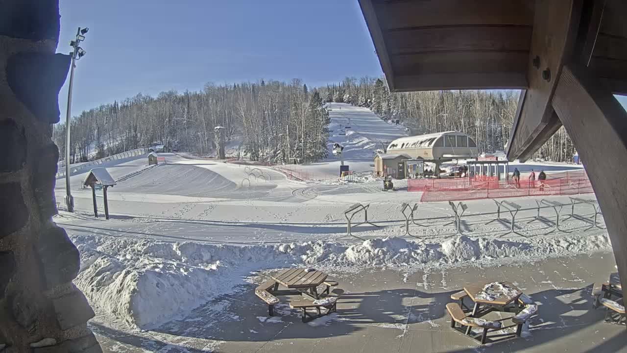 A snowy ski resort is illuminated by floodlights at night, featuring a large lighted Christmas tree structure, ski lifts, and snow-covered picnic tables in the foreground under hazy conditions.