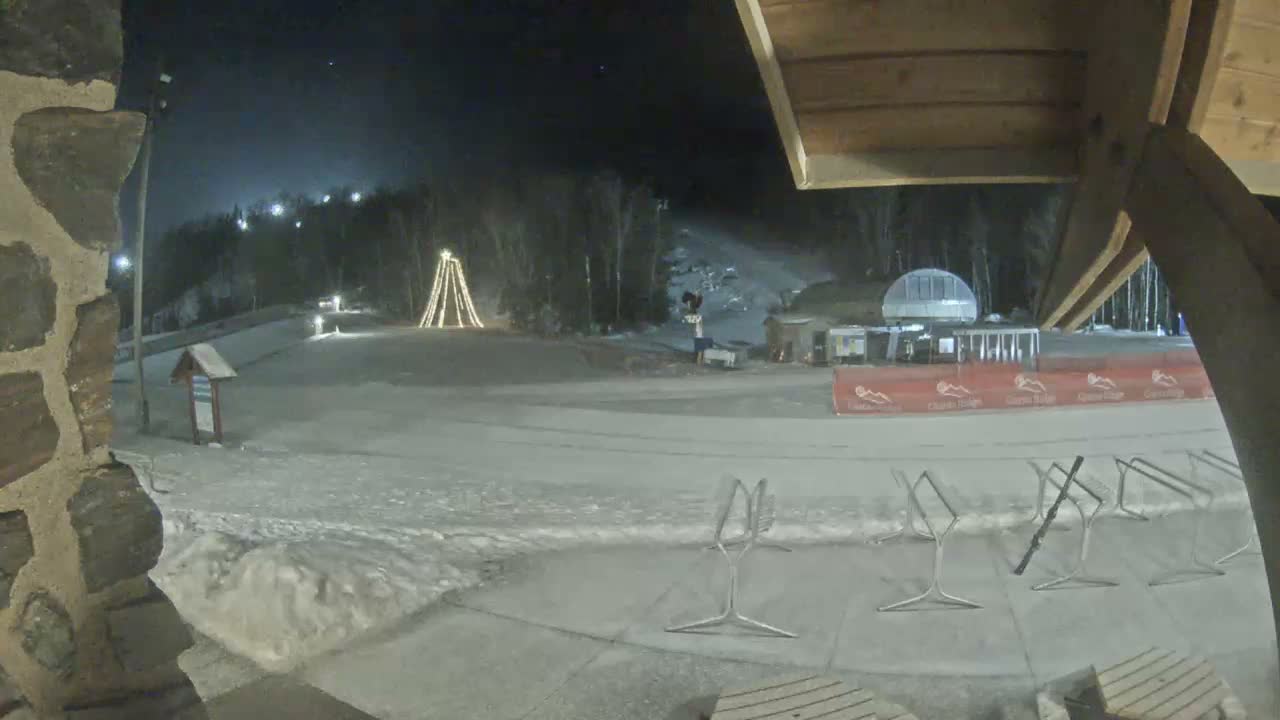 A clear, cold night view of a snow-covered ski resort features a large illuminated structure, a ski lift station, and groomed slopes, seen from under a wooden canopy beside a stone wall.