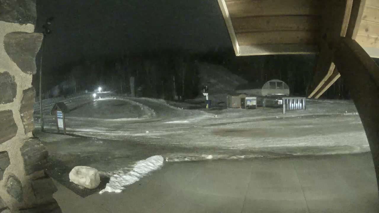 A clear, cold night view of a snow-covered ski resort features a large illuminated structure, a ski lift station, and groomed slopes, seen from under a wooden canopy beside a stone wall.