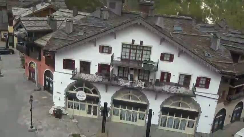 An aerial view shows a cluster of buildings with dark slate roofs, one large white building with balconies and arched storefronts in the center, under an overcast sky.